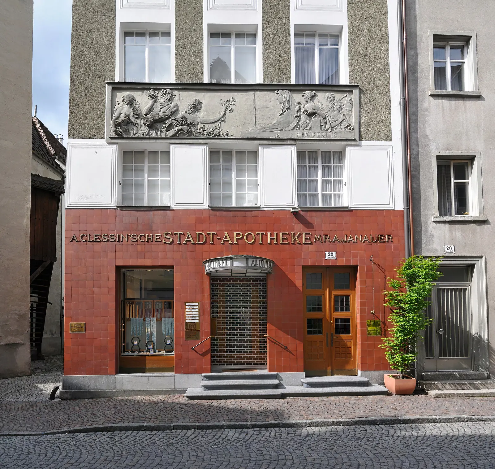 Fassade der Stadt-Apotheke frontal: Relief-Fries über dem ersten Obergeschoss, darunter das rote Erdgeschoss mit goldener Inschrift